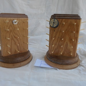 Op de afbeelding: Twee houten spoelhouders met een donkerbruine basis en lichtbruine houten zijden. Elke houder heeft een rij kleine houten pinnen om spoelen garen vast te houden. De ene houder heeft een kleine ronde metalen knop aan de bovenkant en de andere heeft een kleine ronde zwarte spoel garen aan de bovenkant.