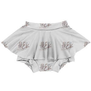 May include: A white baby onesie with a ruffled skirt. The onesie has a repeating pattern of the text "HER GRAPHICS WHOLESALE" in a brown and gray script font.
