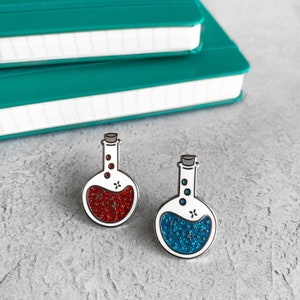 May include: Two enamel pins shaped like glass bottles with cork stoppers. The bottles are filled with glitter, one red and one blue.