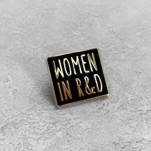 Może przedstawiać: Czarna szpilka emaliowana ze złotym napisem "Women in R&D".
