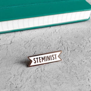 May include: A white enamel pin with a gold border in the shape of a banner. The banner reads "STEMINIST" in black lettering.