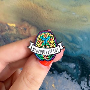 Puede incluir: Un pin de esmalte colorido con un diseño de cerebro con un patrón de arcoíris. El pin tiene una pancarta que dice "Neurodivergent" en texto blanco. El pin está sostenido por una mano con esmalte de uñas rojo.