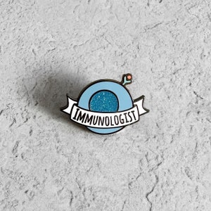 Peut inclure: Épinglette émaillée bleu clair avec un motif circulaire et le mot "IMMUNOLOGIST" sur une bannière. Le centre du cercle a une texture bleu pailleté. Un petit motif rose et rouge se trouve en haut à droite du cercle.