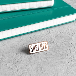 May include: White enamel pin with a gold outline and the text "SHE/HER" in black lettering.