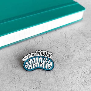 Peut inclure: Une épingle en émail bleu et blanc avec une illustration de dessin animé d'une mitochondrie. L'épingle dit "I've got the power" au-dessus de la mitochondrie.