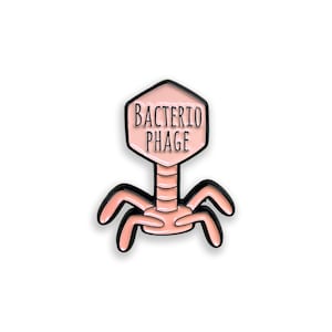 Op de afbeelding: Een lichtroze emaille speld in de vorm van een bacteriofaagvirus met een zeshoekige kop en zes poten. De kop bevat de zwarte tekst BACTERIO PHAGE in een eenvoudig lettertype. Het ontwerp is omlijnd met zwart metaal voor een duidelijk contrast.