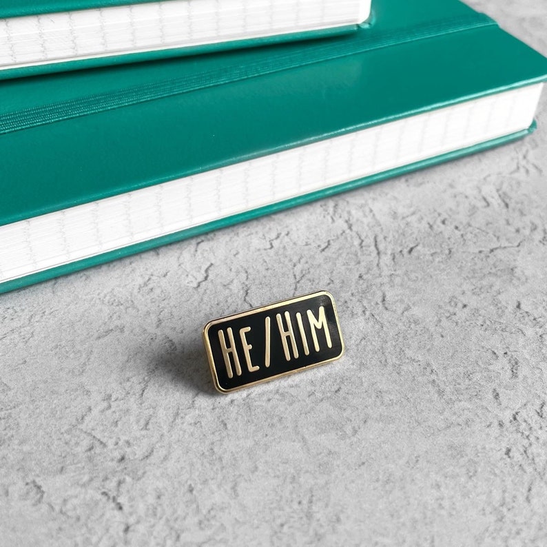 A black enamel pin with gold trim. The pin reads "HE/HIM" in gold lettering.