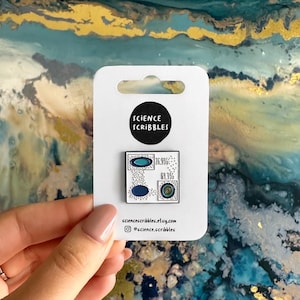 May include: A white enamel pin with a science-themed design. The pin features a black circle with "SCIENCE SCRIBBLES" and a square with blue and white graphics and percentages. The pin is on a white card with a website and social media handle.