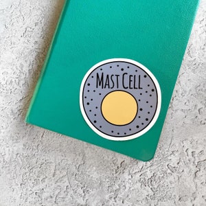 May include: A green notebook with a round sticker that says "Mast Cell" in black text. The sticker has a grey background with a yellow circle in the centre and black dots around the edge.