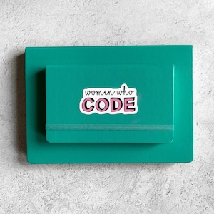 May include: A teal notebook with a pink and white sticker that says "women who code" on it.