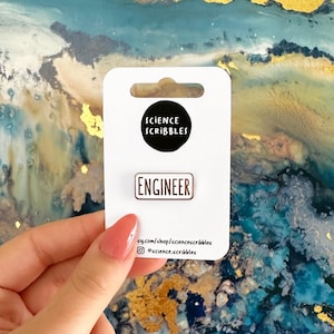 May include: A white card with the text "Science Scribbles" and a gold pin with the word "Engineer" on it. The card has a website and social media handle printed on it.