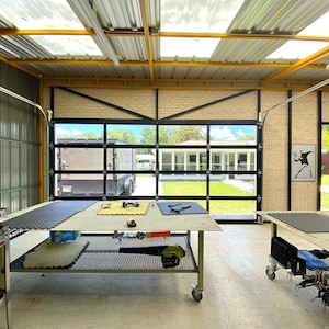 Op de afbeelding: Een werkplaats met een grote garagedeur die openstaat, met uitzicht op een achtertuin met een huis en groen gras. Er zijn twee werkbanken met zwarte rubberen matten erop, en verschillende gereedschappen en apparatuur zijn verspreid over de ruimte. Het plafond heeft gele balken en dakramen.