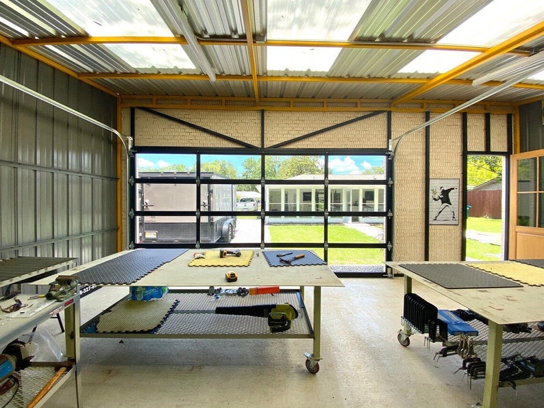 Op de afbeelding: Een werkplaats met een grote garagedeur die openstaat, met uitzicht op een achtertuin met een huis en groen gras. Er zijn twee werkbanken met zwarte rubberen matten erop, en verschillende gereedschappen en apparatuur zijn verspreid over de ruimte. Het plafond heeft gele balken en dakramen.