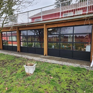Op de afbeelding: Drie grote zwarte garagedeuren met glazen panelen, in een houten frame geplaatst. De deuren staan open en geven uitzicht op een tuin en bomen.