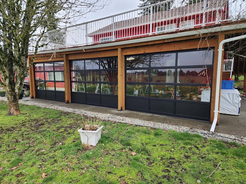 Op de afbeelding: Drie grote zwarte garagedeuren met glazen panelen, in een houten frame geplaatst. De deuren staan open en geven uitzicht op een tuin en bomen.