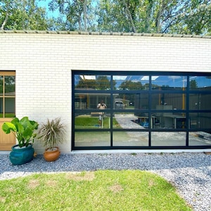 Op de afbeelding: Een garage van witte bakstenen met een zwarte glazen garagedeur en een houten deur met glazen panelen. Er staan twee planten in potten voor de garage.