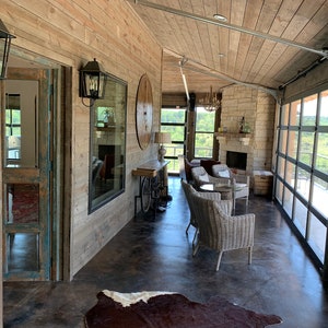Op de afbeelding: Een rustieke woonkamer met een open haard, een groot raam en een glazen garagedeur. De kamer is ingericht met rieten stoelen en een koeienhuidkleed.