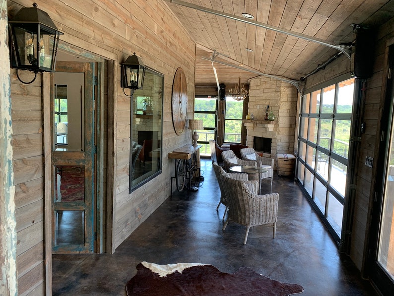 Op de afbeelding: Een rustieke woonkamer met een open haard, een groot raam en een glazen garagedeur. De kamer is ingericht met rieten stoelen en een koeienhuidkleed.