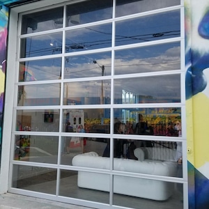 Op de afbeelding: Een witte, moderne bank staat voor een grote glazen garagedeur. De deur is gemaakt van meerdere ruiten en heeft een zilveren frame. De deur staat open en de bank is zichtbaar door het glas. De bank is modern ontworpen met een laag profiel en een witte lederen bekleding.