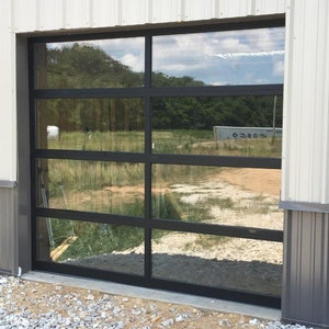 Op de afbeelding: Een glazen garagedeur met zwart frame en vijf horizontale panelen. De deur is gesloten en reflecteert het omringende landschap.