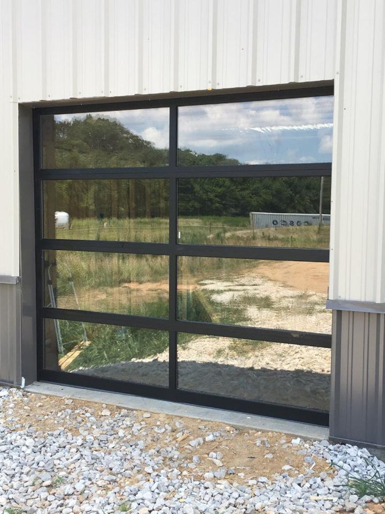 Op de afbeelding: Een glazen garagedeur met zwart frame en vijf horizontale panelen. De deur is gesloten en reflecteert het omringende landschap.