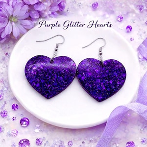 May include: A pair of heart-shaped earrings with a glittery purple finish. The earrings hang from silver-colored hooks. The words "Purple Glitter Hearts" are written above the earrings. The earrings are displayed on a white plate.