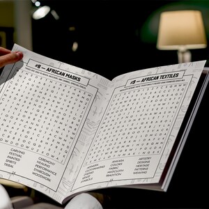 K&ouml;nnte beinhalten: Ein offenes Buch mit den &Uuml;berschriften "#8 - Afrikanische Masken" und "#9 - Afrikanische Textilien." Die Seiten zeigen Wortsuchr&auml;tsel und verwandte Schl&uuml;sselw&ouml;rter wie "Schnitzen", "Farben" und "Weben". Das Buch wird von einer Hand gehalten.