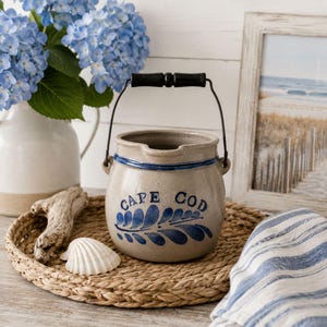May include: A beige stoneware crock featuring a blue painted leaf design and the text CAPE COD. The crock has a dark metal handle with a black wooden grip, a blue rim, and a small pouring spout. It sits on a round woven seagrass tray.