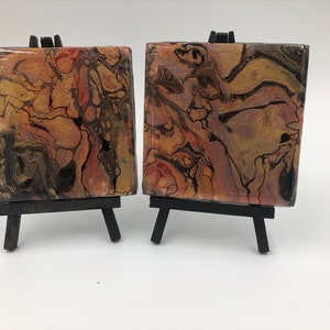 May include: Two small abstract paintings on canvases with black, brown, gold, and pink swirling patterns. The canvases are displayed on small black easels.