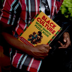K&ouml;nnte beinhalten: Eine Person h&auml;lt ein gelbes "Black Culture Word Search Puzzle Book". Das Buchcover zeigt Portr&auml;ts und roten Text. Der Titel ist in gro&szlig;en roten Buchstaben gehalten. Das Buch wird vor einem rot-schwarz karierten Hemd gehalten.