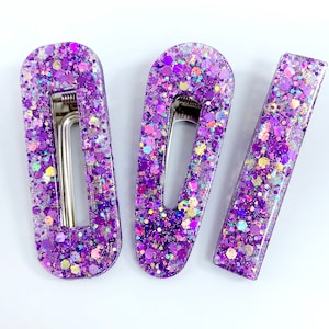May include: Three purple glitter hair clips. The clips are oval shaped with a metal bar in the center. The glitter is iridescent and has a variety of colors.