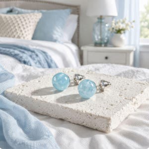 May include: A pair of light blue, round stud earrings with silver posts. The earrings are displayed on a textured, off-white surface, with a soft blue scarf and a bed in the background. The earrings have a marbled appearance.
