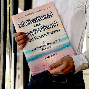 Może przedstawiać: Osoba trzyma książkę zatytułowaną "Motivational and Inspirational Word Search Puzzles". Okładka przedstawia gradient r&oacute;żu, błękitu i bieli z tytułem w kolorze czarnym. Książka opisana jest jako "for Confidence, Strength & Success".