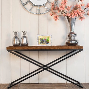 Puede incluir: Una mesa consola de madera con un marco de metal negro y una tapa de madera blanca desgastada. La mesa est&aacute; decorada con dos figuras de pi&ntilde;a plateadas y una impresi&oacute;n enmarcada de una escena de playa con el texto "Welcome Back" en ella.