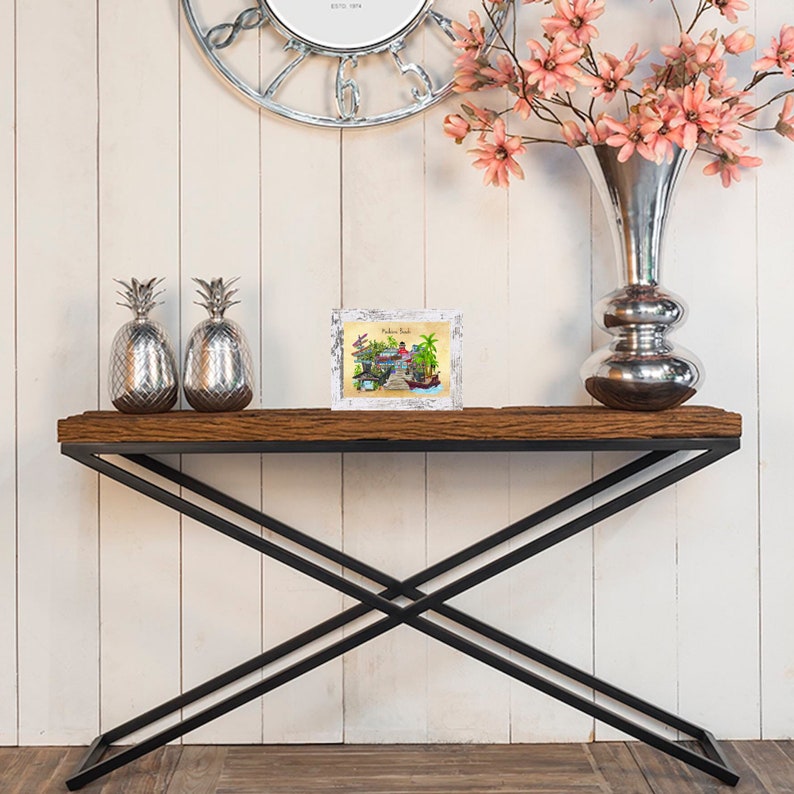 Puede incluir: Una mesa consola de madera con un marco de metal negro y una tapa de madera blanca desgastada. La mesa est&aacute; decorada con dos figuras de pi&ntilde;a plateadas y una impresi&oacute;n enmarcada de una escena de playa con el texto "Welcome Back" en ella.
