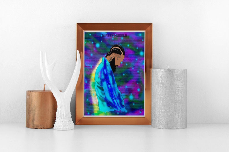 May include: Framed art print featuring a stylized illustration of a person with a beard, wearing a blue jacket, set against a vibrant, colorful background. The text "Nipsey Hussle" is at the top. The artwork is displayed on a white surface.