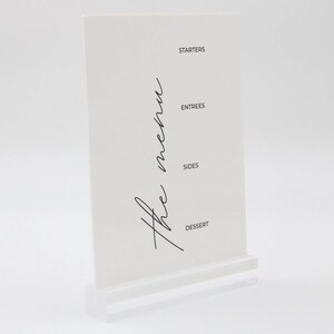 May include: White acrylic menu stand with the words "The Menu" written in black cursive script. The stand has four sections labeled "Starters", "Entrees", "Sides", and "Dessert".