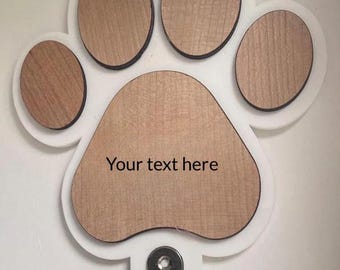 May include: A paw-shaped wall hook with a wooden grain design. The paw print is outlined in white and has a black border. The text "Your text here" is printed on the paw. A green and black dog leash hangs from the metal hooks.