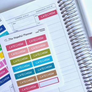 May include: A planner with a spiral binding is open to a page with the text "Wednesday, July 28" and "Catechism." A sheet of colorful "Catechism" labels in various colors, including pink, green, and blue, is next to the planner.