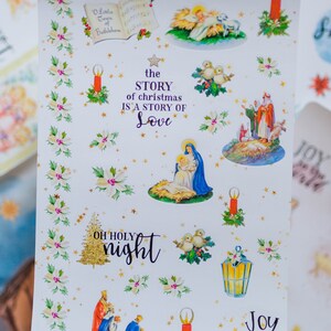 May include: A sheet of Christmas stickers featuring a nativity scene, candles, doves, a star, and a Christmas tree. The stickers are on a white background with gold stars and holly leaves. The text "The Hopeful Planner" is at the top of the sheet. The text "the STORY of christmas IS A STORY OF Love" is in the center of the sheet. The text "OH HOLY night" is at the bottom left of the sheet. The text "Joy to the world" is at the bottom right of the sheet.
