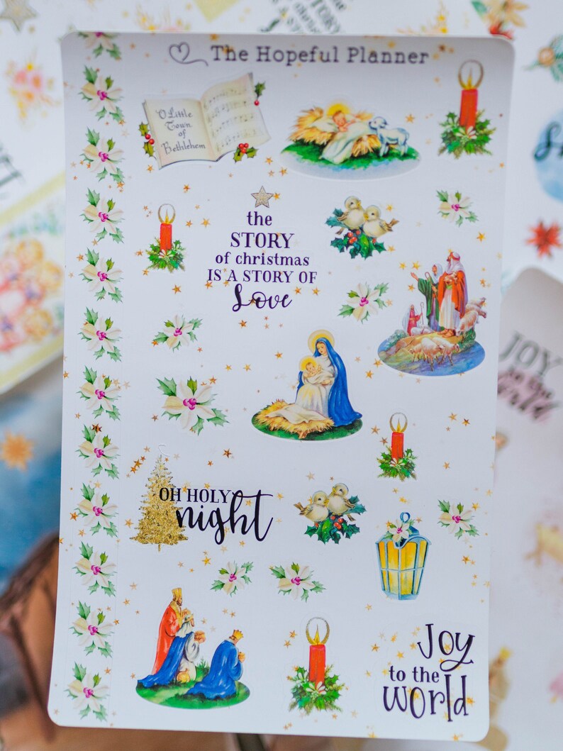 May include: A sheet of Christmas stickers featuring a nativity scene, candles, doves, a star, and a Christmas tree. The stickers are on a white background with gold stars and holly leaves. The text "The Hopeful Planner" is at the top of the sheet. The text "the STORY of christmas IS A STORY OF Love" is in the center of the sheet. The text "OH HOLY night" is at the bottom left of the sheet. The text "Joy to the world" is at the bottom right of the sheet.