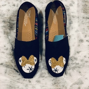Op de afbeelding: Paar zwarte instappers met een Minnie Mouse-ontwerp. Elke schoen heeft een gouden glitterstrik en een witte cirkel met sneeuwvlokaccenten. De binnenkant heeft een patroon en de merknaam "TOMS".