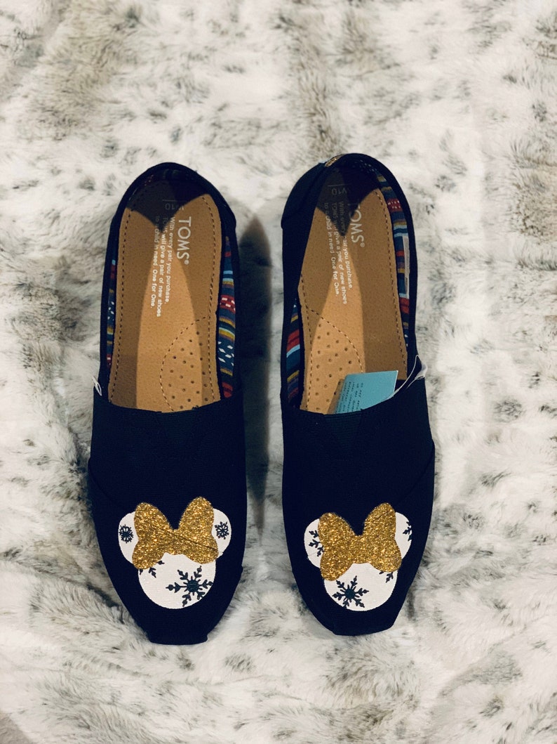 Op de afbeelding: Paar zwarte instappers met een Minnie Mouse-ontwerp. Elke schoen heeft een gouden glitterstrik en een witte cirkel met sneeuwvlokaccenten. De binnenkant heeft een patroon en de merknaam "TOMS".