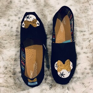 Op de afbeelding: Twee zwarte TOMS instappers, elk versierd met een wit en goud glitter Minnie Mouse ontwerp. De schoenen hebben een strik en sneeuwvlok details. De binnenkant heeft een patroon voering en het TOMS logo.