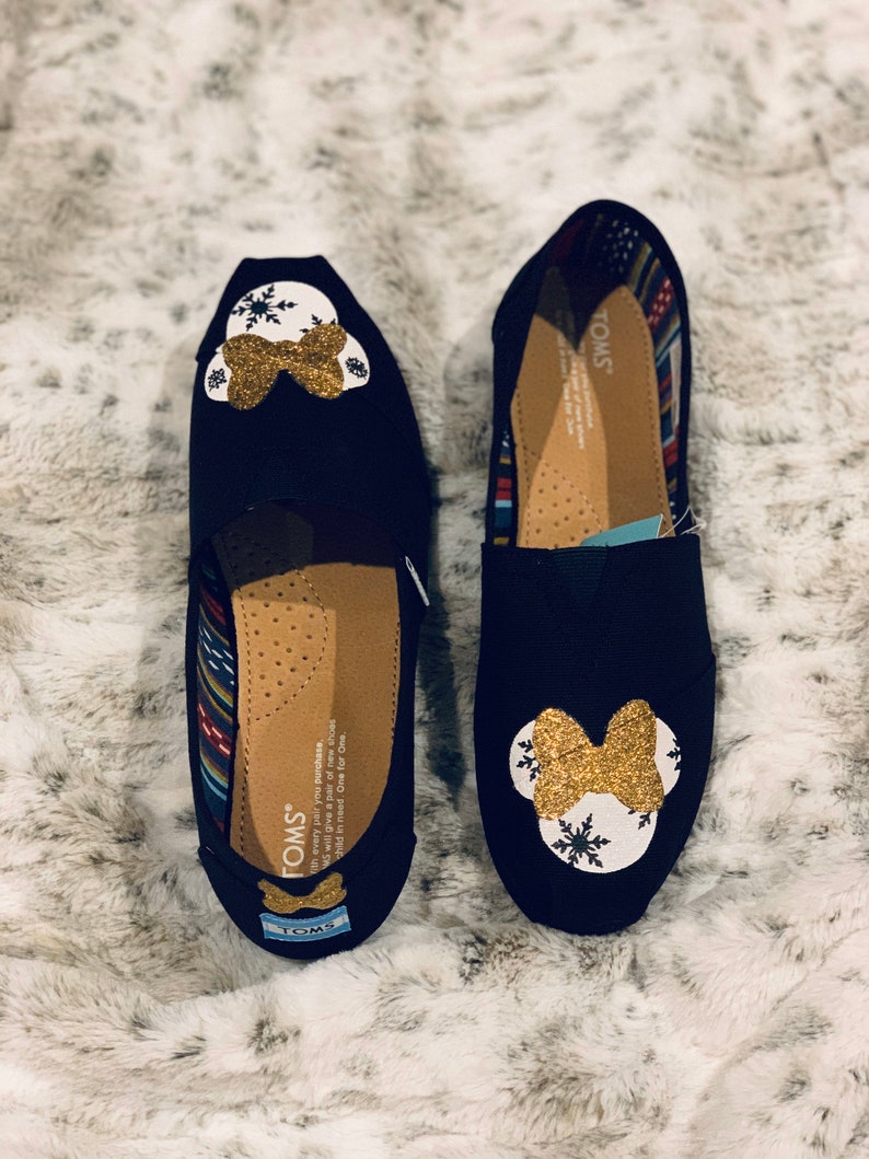 Op de afbeelding: Twee zwarte TOMS instappers, elk versierd met een wit en goud glitter Minnie Mouse ontwerp. De schoenen hebben een strik en sneeuwvlok details. De binnenkant heeft een patroon voering en het TOMS logo.