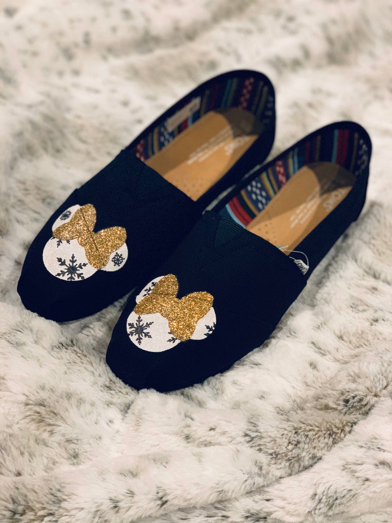 Op de afbeelding: Een paar zwarte instappers met een gouden glitterstrik en een wit sneeuwvlokontwerp. De schoenen hebben een kleurrijk patroon aan de binnenkant en worden gepresenteerd op een zacht, wit oppervlak. Schoenmaat 38.
