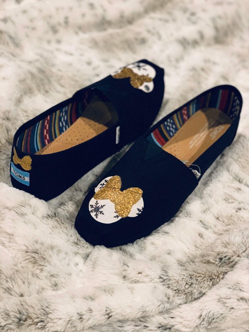 Op de afbeelding: Een paar zwarte instappers met een goudkleurig glitterontwerp en een witte sneeuwvlok. De schoenen hebben een patroon aan de binnenkant en de merknaam "TOMS" op de hiel.
