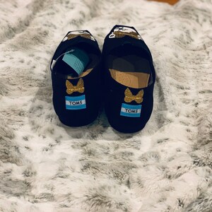 Op de afbeelding: Een paar zwarte TOMS instappers met gouden glitter vlinder accenten en het TOMS logo op de hiel. De schoenen hebben een cartoon karakter ontwerp op de bovenkant en worden weergegeven op een zacht, wit en grijs oppervlak.