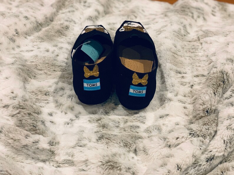 Op de afbeelding: Een paar zwarte TOMS instappers met gouden glitter vlinder accenten en het TOMS logo op de hiel. De schoenen hebben een cartoon karakter ontwerp op de bovenkant en worden weergegeven op een zacht, wit en grijs oppervlak.