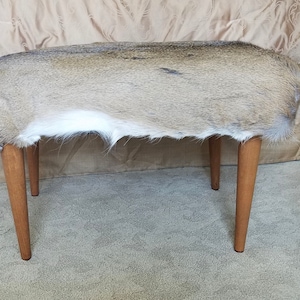 May include: A small, upholstered bench with a natural deer hide top. The hide is a mix of tan, brown, and white, with a soft, furry texture. The bench has four wooden legs, each angled slightly outward. The legs are a light brown color.